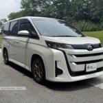 Front three-quarter view of a white 2024 Toyota Noah Hybrid 1.8A X with bronze sports rims.